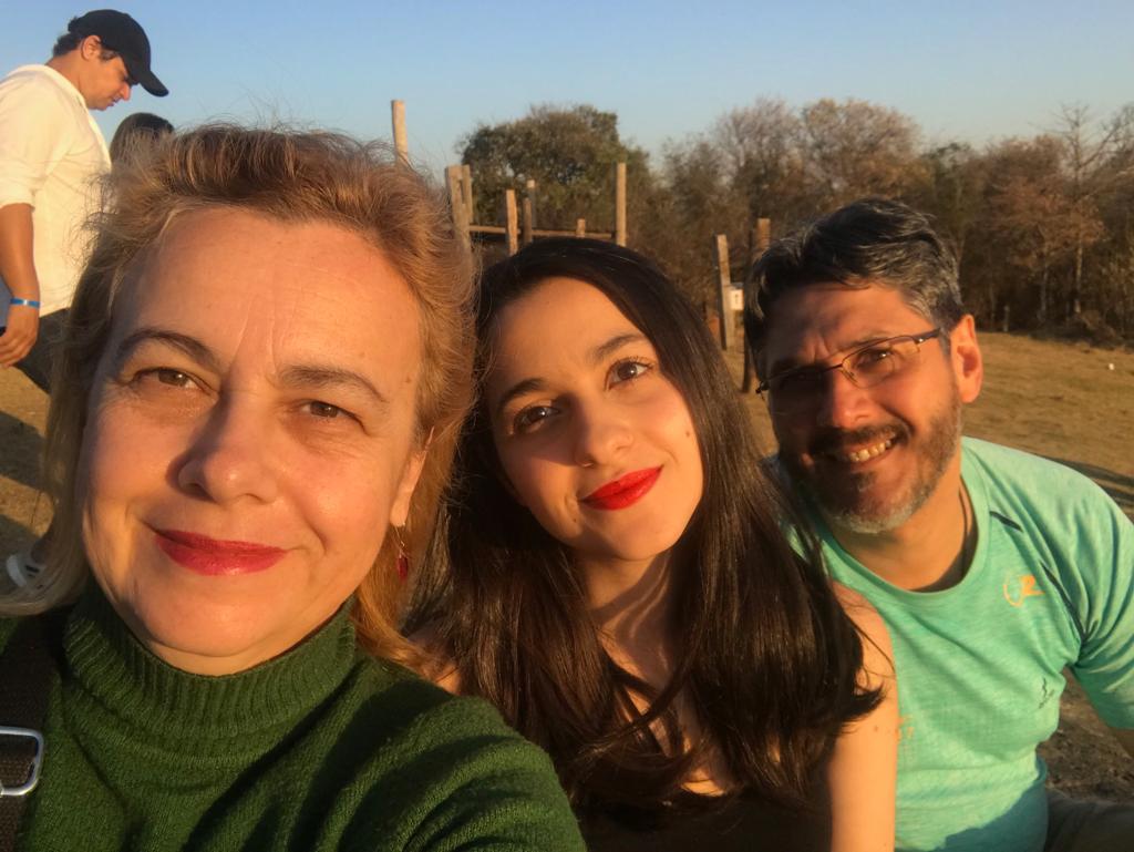 Sentados no gramado de um parque, perto um do outro, Celeste, loira e de blusa verde, sorri ao lado da filha Maria Luiza, de cabelos castanhos e batom vermelho, e do marido Edson, grisalho, de óculos, barba e camiseta azul clara,que também sorri de óculos e